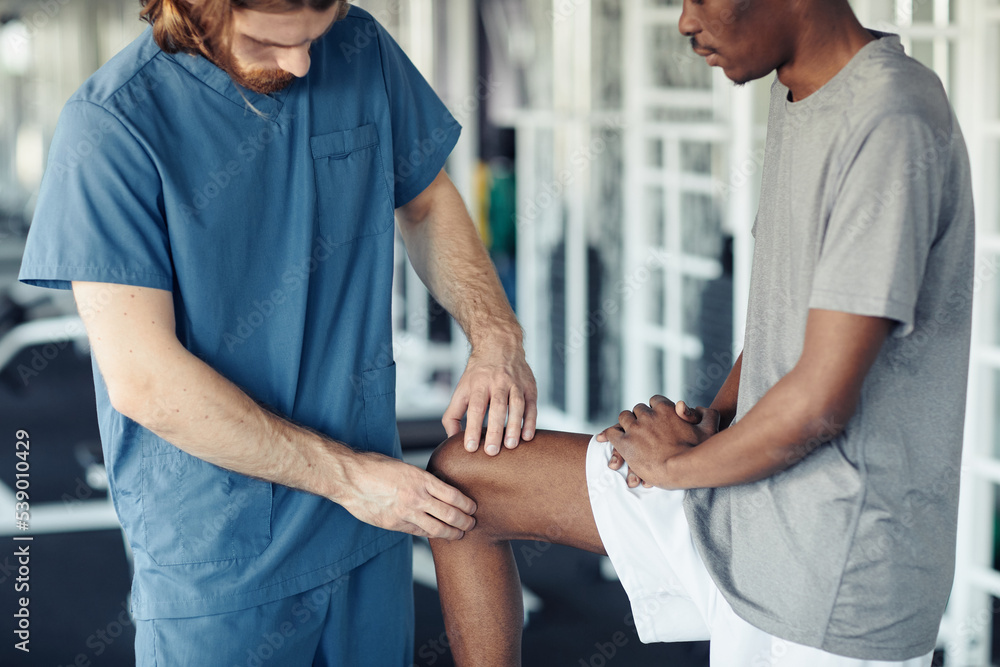 Fototapeta premium Close-up of doctor in uniform examining the pain leg of African patient in gym