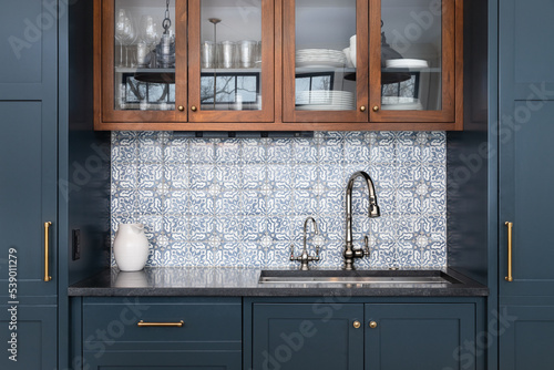 Fotótapéta A kitchen sink with a beautiful pattern tiled backsplash with a chrome faucet, black granite countertops, and surrounded by blue and wood cabinets