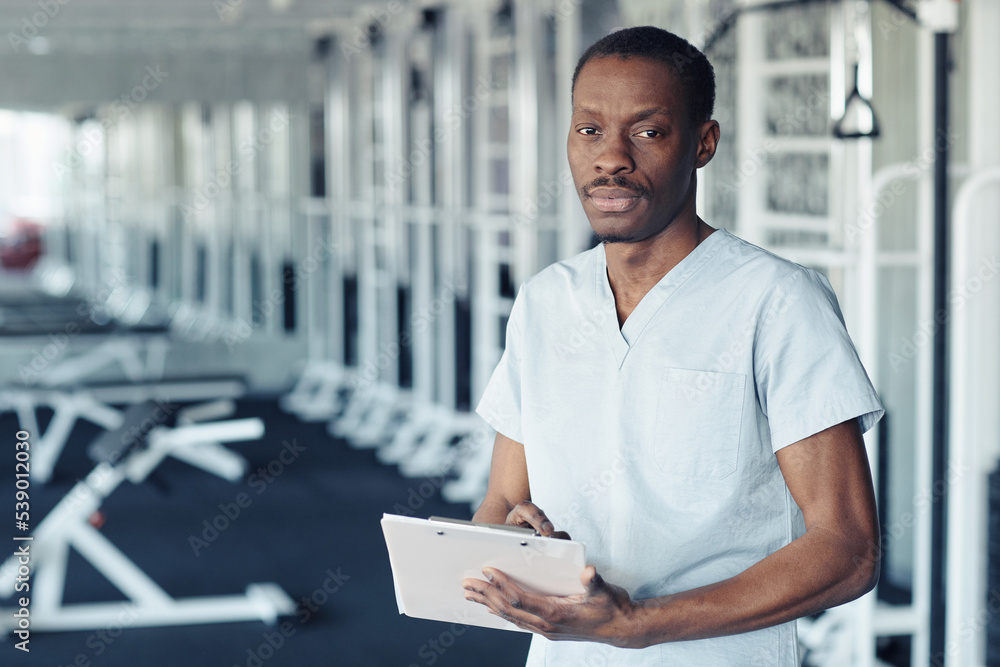 Portrait of African doctor in uniform filling patient medical card while standing in gym
