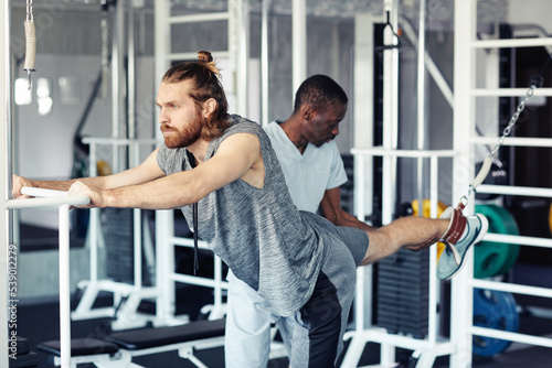 Young patient stretching his injury leg on sport equipment in gym with therapist helping him