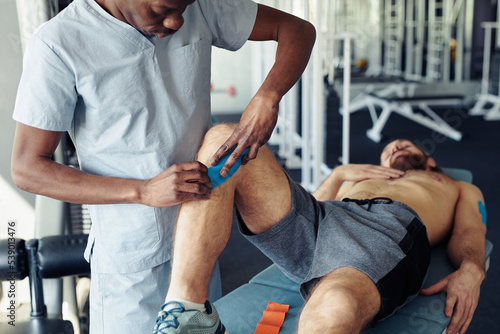 Fototapeta Naklejka Na Ścianę i Meble -  African young therapist applying medical tapes on legs of patient while he lying on couch before sport training