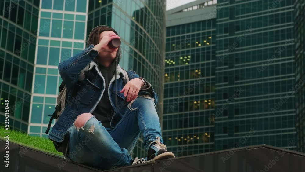 Young man with dreadlocks drinking coffee on background of skyscrapers. Relaxed hipster enjoying hot drink, sitting on stairway in downtown. Young man with dreadlocks drinking coffee on background of skyscrapers. Relaxed hipster enjoying hot drink, sitting on stairway in downtown.