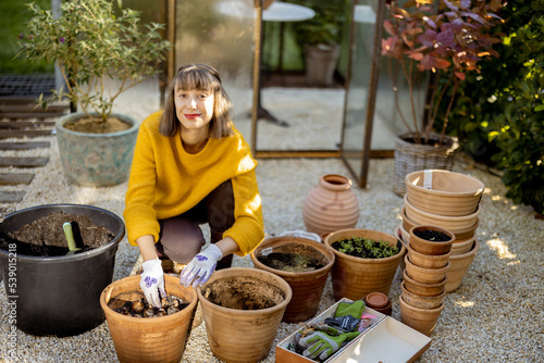 Portrait of a young woman planting flower bulbs in clay jugs sitting in front of glass orangery at garden. Florist gardening at beautiful backyard on sunny morning