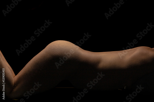 Artistic nude male fitness model with lighting on bare skin from back to thighs against black background