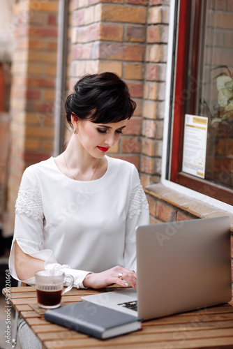 Young businesswoman working on laptop at cafe on terrace in urban city, stylish outfit with white blouse and skirt. Coffee break, business concept. Working outdoors. Remote job.