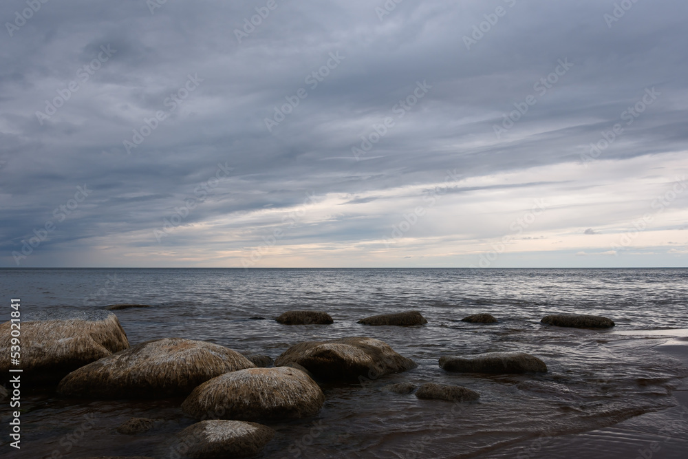 Baltic Sea before sunset. Evening cloudy sky over the coast of Narva Bay. The calm sea washes coastal granite stones. Northern seascape with light dramatic notes. Baltic Sea before sunset. Evening cloudy sky over the coast of Narva Bay. The calm sea washes coastal granite stones. Northern seascape with light dramatic notes.