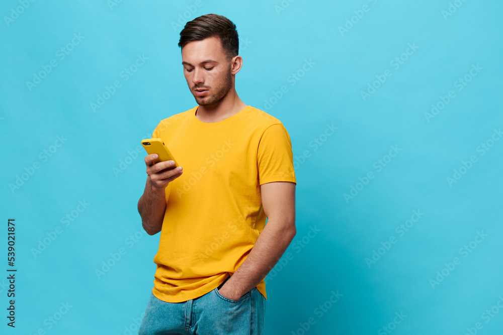 Thoughtful concentrated tanned handsome man in yellow t-shirt read news online shopping using phone posing isolated on blue studio background. Copy space Banner Mockup. Online communication concept Thoughtful concentrated tanned handsome man in yellow t-shirt read news online shopping using phone posing isolated on blue studio background. Copy space Banner Mockup. Online communication concept