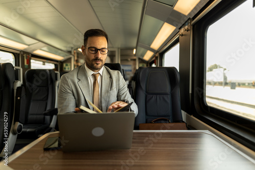 Formal wearing business man traveling to work by train. Business man is working while traveling, using laptop, mobile phone, and taking notes. Business man planning goals and meetings © Nemanja