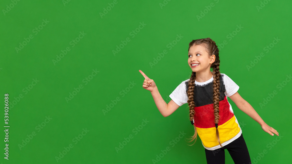 A little girl with a German flag on her T-shirt points to your advertisement on a green isolated background. German language courses for schoolchildren. Copy space. A little girl with a German flag on her T-shirt points to your advertisement on a green isolated background. German language courses for schoolchildren. Copy space.
