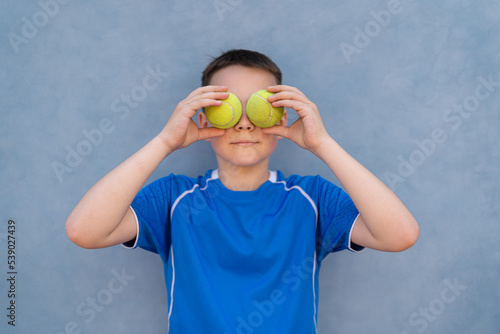 Portrait of a young boy holding tennis balls over his eyes on blue background. Sports concept © diignat