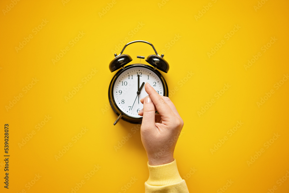 Daylight saving day. Fall Back. Black Alarm clock and female hands on yellow background. Daylight saving time end - obrazy, fototapety, plakaty 