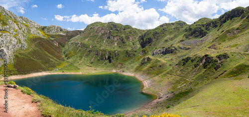 Fototapeta Naklejka Na Ścianę i Meble -  Vistas del paisaje asturiano del Lago de La Cueva por un sendero rodeado de naturaleza, montañas verdes, flores moradas cielo azul y nubes blancas en verano de 2021, España.
