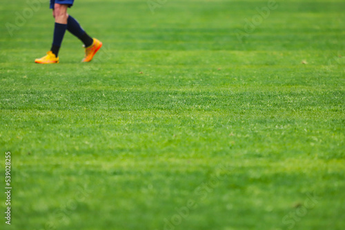 Green grass of a soccer field with selective focus in the center of the image, above the image some feet with cleats.
