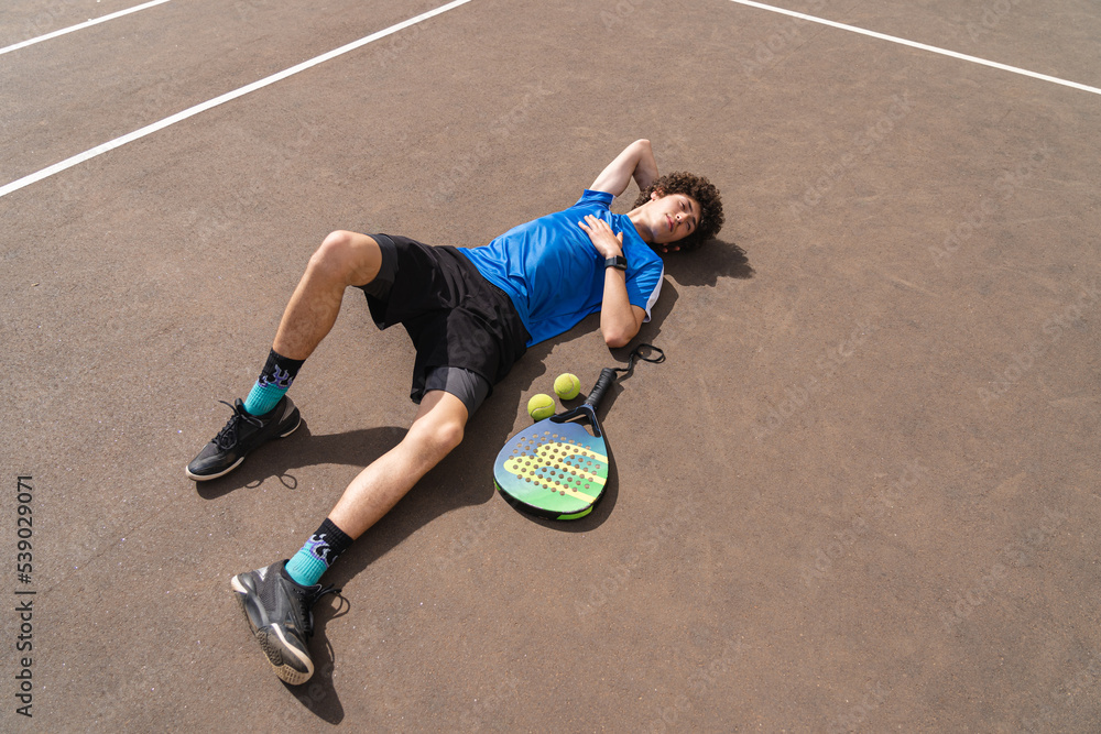 Portrait of sporty young man with curly hair lying on the padel outdoor court with a racket and a ball. Spots concept, healthy lifestyle. Copy space on the ground.