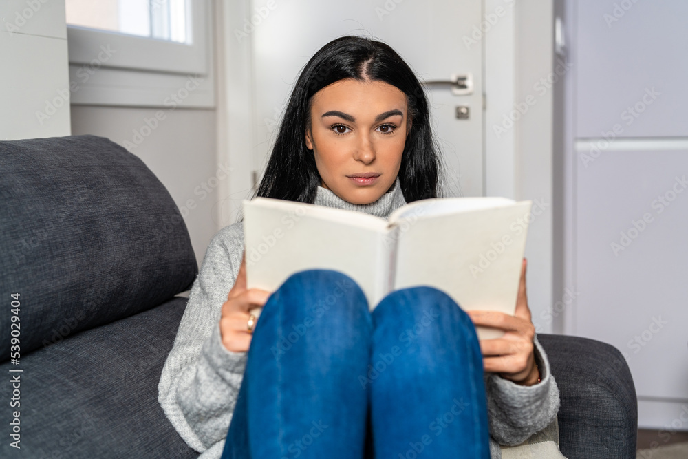 Young woman with long dark hair lying on the sofa while reading a book Young woman with long dark hair lying on the sofa while reading a book