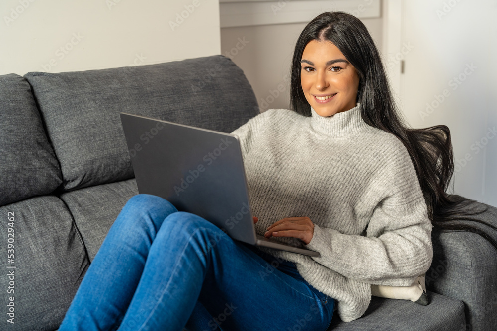 Naklejka premium Young woman with long brunette hair sitting on sofa looking at camera with laptop on her legs