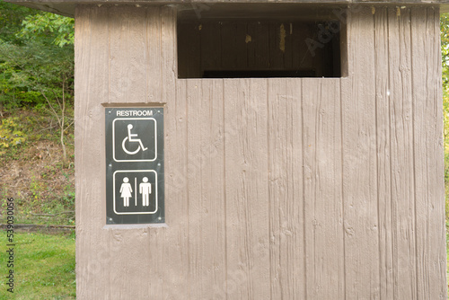 Outhouse vault toilet with accessible restroom and gender signs in public park picnic area parking lot basically just a hole in the ground with a toilet seat on top, the human waste emits methane