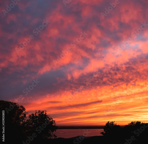 Attractive colours and textures of clouds at sunset over a bay of water with silhouetted trees in the foreground.