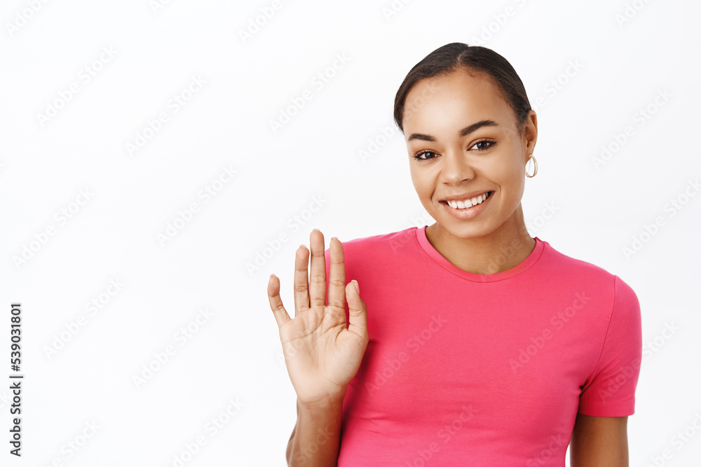Close up of smiling black woman says hello and waves hand, greets you, stands in pink tshirt over white background Close up of smiling black woman says hello and waves hand, greets you, stands in pink tshirt over white background