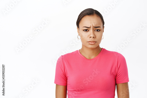 Close up of girl looks concerned, frowns with worried face expression, stands in pink tshirt over white background