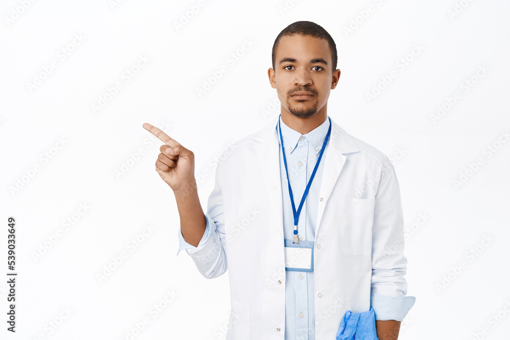 Doctor in white coat pointing finger left. Young intern in clinic showing way, demonstrating promo, standing over white background Doctor in white coat pointing finger left. Young intern in clinic showing way, demonstrating promo, standing over white background
