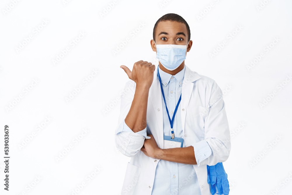 Surprised doctor in medical mask from covid, pointing left, looking amazed, standing over white background Surprised doctor in medical mask from covid, pointing left, looking amazed, standing over white background