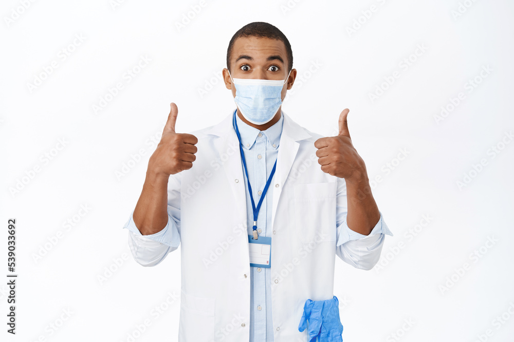 Enthusiastic young doctor in medical mask, shows thumbs up, looks impressed, recommends smth awesome, stands over white background Enthusiastic young doctor in medical mask, shows thumbs up, looks impressed, recommends smth awesome, stands over white background