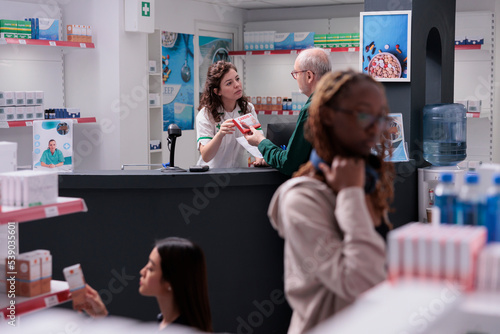 Helpful pharmacy worker discussing about medication treatment with elderly client, explaining pills leaflet during medical consultation. Customer buying supplements, vitamin to cure disease