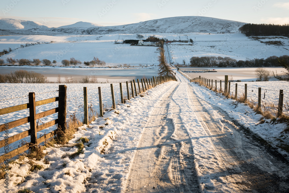 Tyre tracks on a road flanked by wooden fences covered in snow leading to the hills in the distance under a blue sky in a sunny day in the Pentland Hills Regional Park in Edinburgh, Scotland, UK