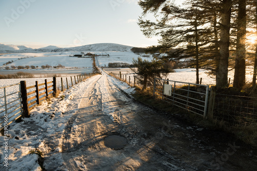 A road flanked by wooden fences covered in snow leading to the hills in the distance under a blue sky in a sunny day in the Pentland Hills Regional Park in Edinburgh, Scotland, UK