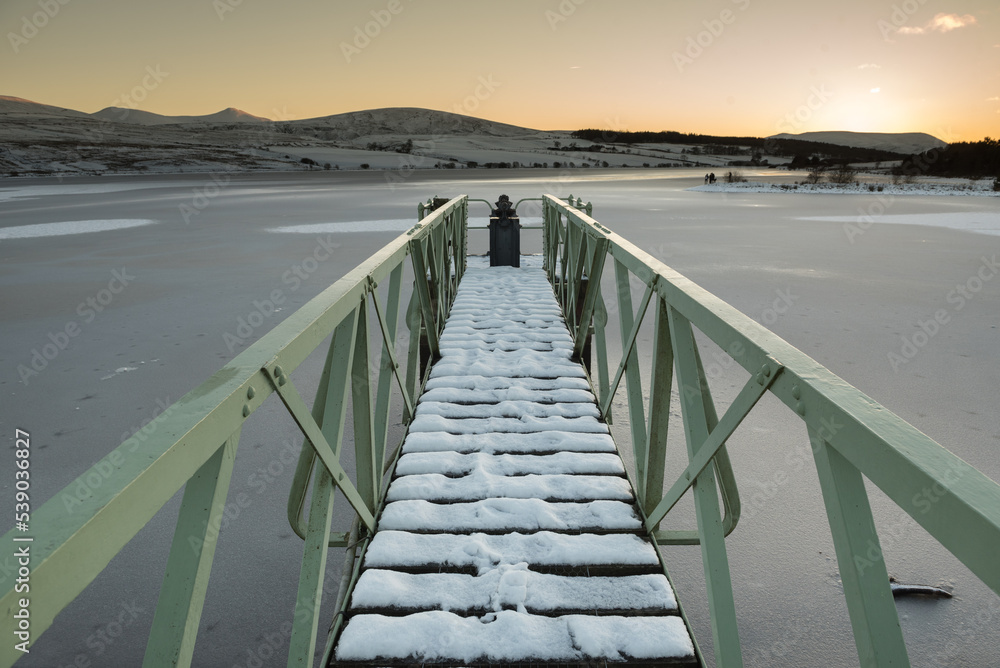 View of a metallic bridge partially covered in snow overlooking a frozen reservoir during sunset in a cold Winter day in the Pentland Hills Regional Park in Edinburgh, Scotland, UK