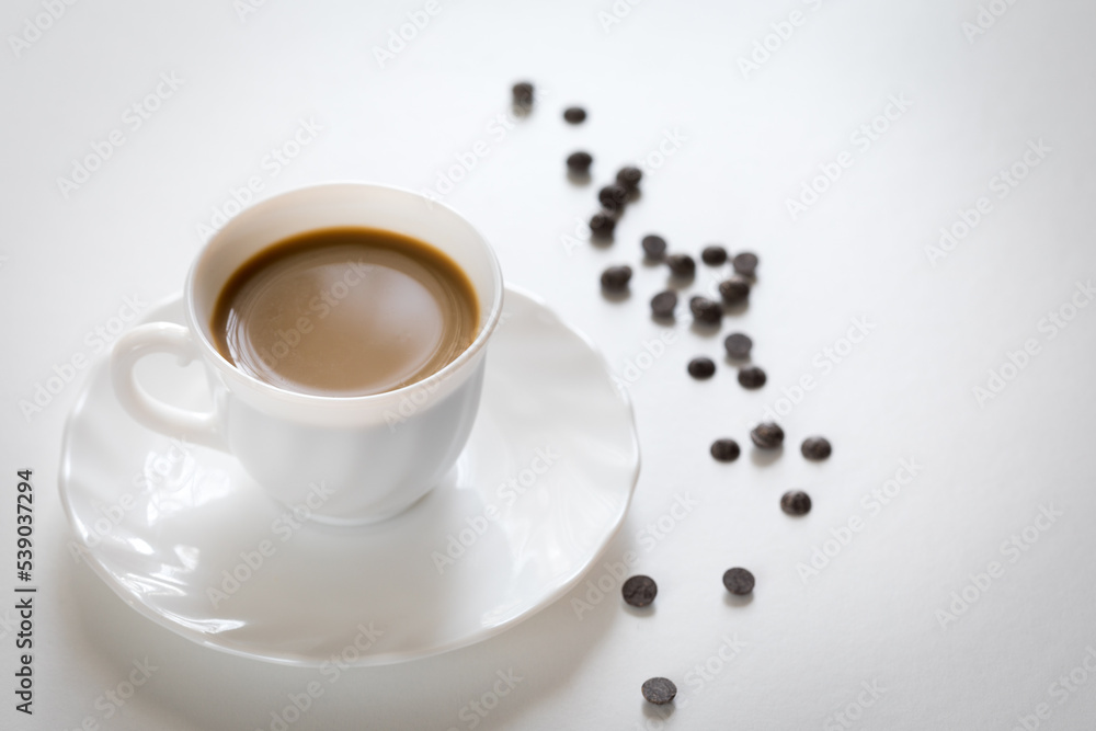 Fototapeta premium Tazza bianca con caffè e piattino con chicchi di cioccolato su tavolo bianco