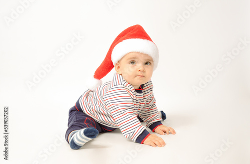 Adorable child is sitting on floor, wearing red Christmas cap, isolated over white background.