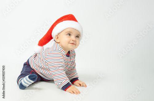 Adorable child is sitting on floor, wearing red Christmas cap, isolated over white background.
