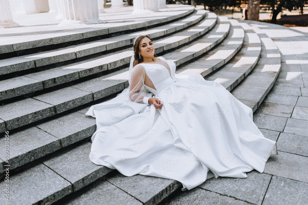 A beautiful bride in a white long dress is sitting on the steps in the park against the backdrop of ancient architecture. Wedding photography, portrait. A beautiful bride in a white long dress is sitting on the steps in the park against the backdrop of ancient architecture. Wedding photography, portrait.
