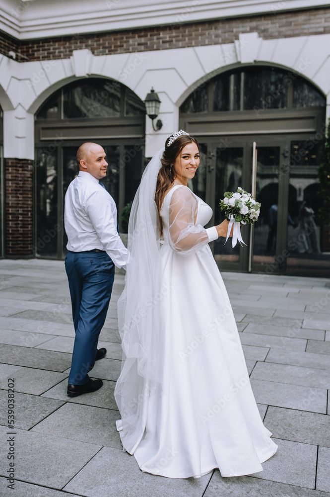 A stylish groom in a blue suit and a beautiful bride in a white dress walk holding hands in the city, in the park. Wedding photography, portrait.