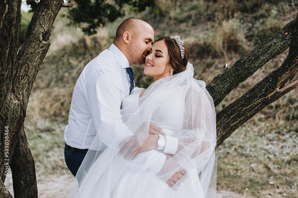 A stylish adult groom and a beautiful, young, smiling bride in a white dress are hugging, standing in nature near a tree. Close-up wedding photography, portrait. A stylish adult groom and a beautiful, young, smiling bride in a white dress are hugging, standing in nature near a tree. Close-up wedding photography, portrait.