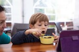 Little boy sit on chair at table and with interest watch cartoon on tablet computer with mother. Kid of kindergarten age open mouth to eat chocolate balls with milk. Ready breakfast, gadget addiction