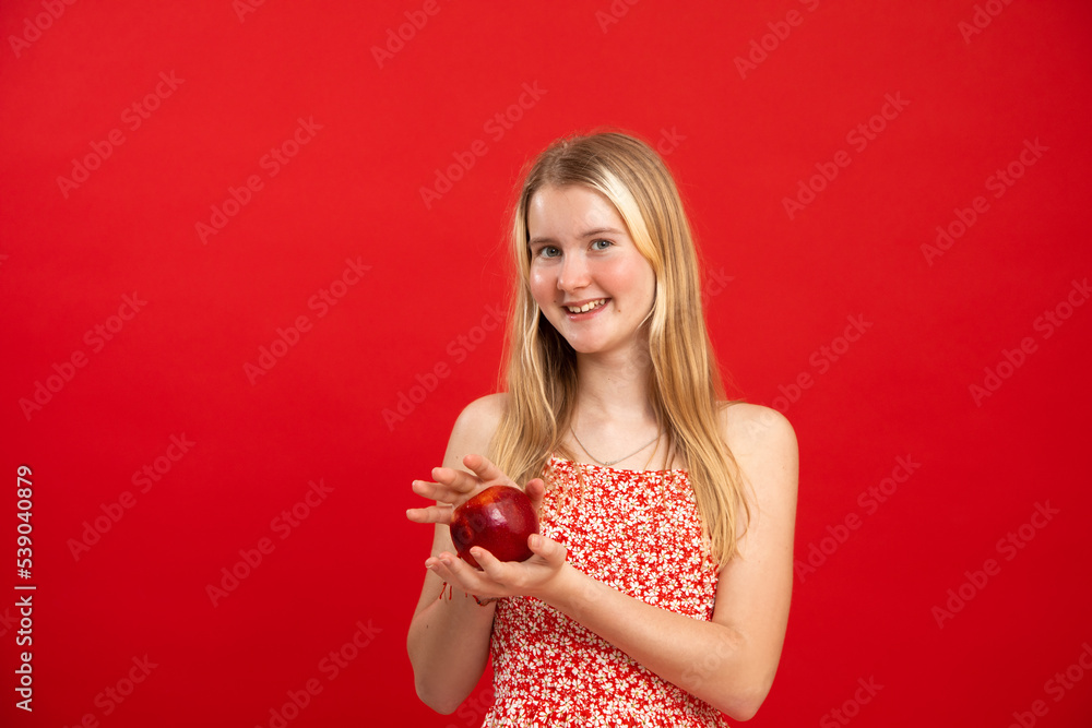 Blonde haired teen girl stand in floral print dress and hold ripe fresh apple on empty red background, free copy space. Portrait of happy school aged child with healthy food. Nutrition, diet, fruits Blonde haired teen girl stand in floral print dress and hold ripe fresh apple on empty red background, free copy space. Portrait of happy school aged child with healthy food. Nutrition, diet, fruits