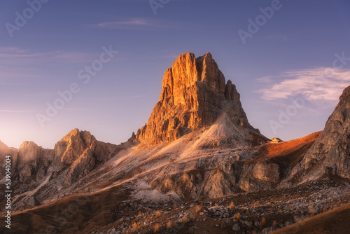 Rocky mountains at colorful sunset in autumn. Mountain pass and beautiful purple sky at dusk. Amazing landscape with rocks, mountain peaks, stones, trails, buildings, trees on hills. Dolomites, Italy