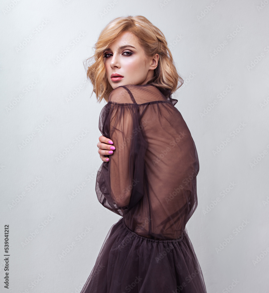 Fototapeta premium Alluring blond beautiful woman with curly short hair style posing in fashion black dress on blue background. Closeup studio portrait