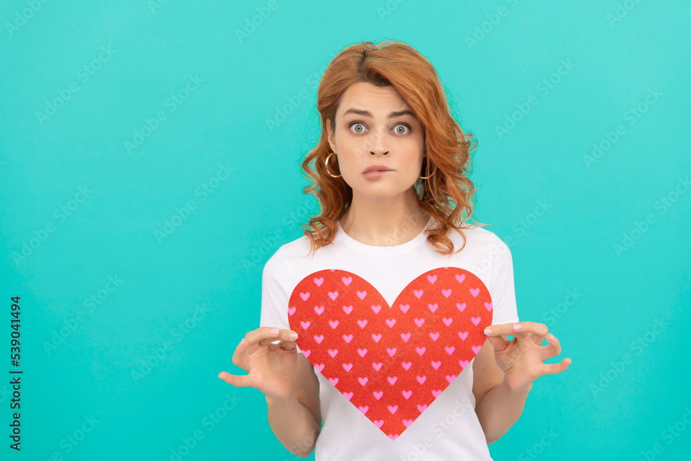 confused redhead woman hold red heart on blue background confused redhead woman hold red heart on blue background