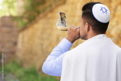 Jewish man in kippah and tallit blowing shofar outdoors. Rosh Hashanah celebration photo