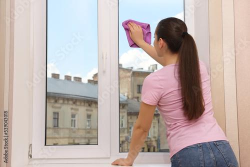 Young woman cleaning window glass with rag at home, back view