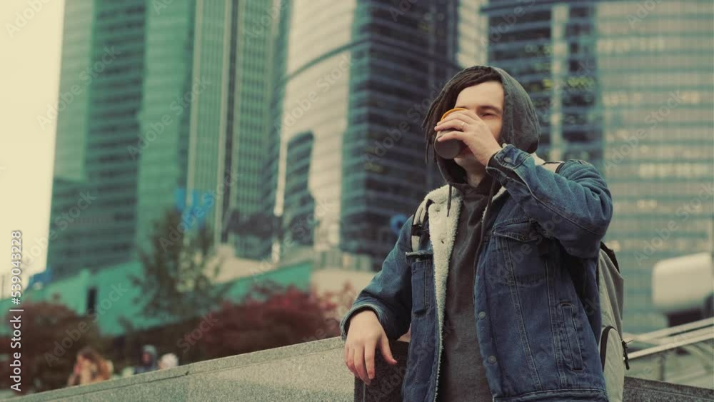 Young man with dreadlocks drinking coffee from cup, standing in background of skyscrapers. Male enjoying hot drink in downtown. Young man with dreadlocks drinking coffee from cup, standing in background of skyscrapers. Male enjoying hot drink in downtown.