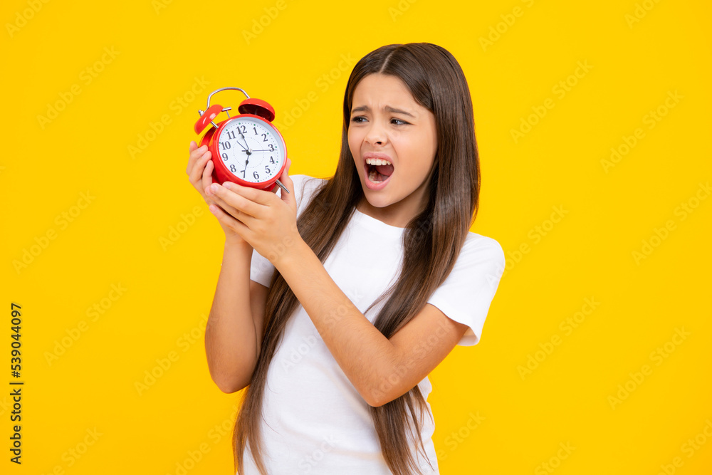 Teen student girl hold clock isolated on yellow background. Time to school. Teenager child with alarm clock showing time, late awakening. Angry face, upset emotions of teenager girl. Teen student girl hold clock isolated on yellow background. Time to school. Teenager child with alarm clock showing time, late awakening. Angry face, upset emotions of teenager girl.