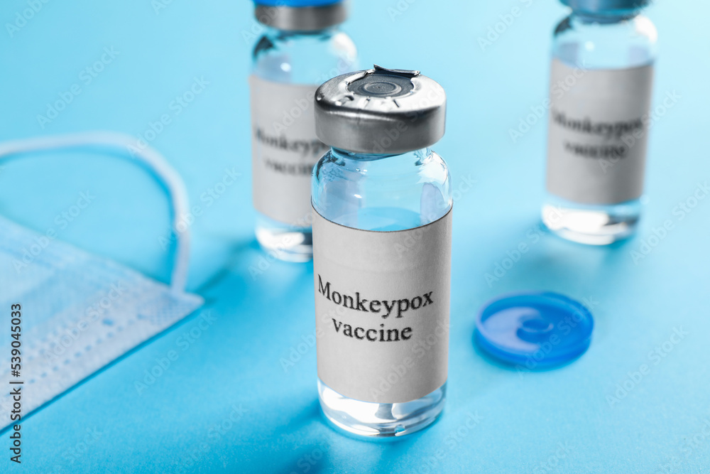 Monkeypox vaccine in glass vials and medical mask on light blue background Monkeypox vaccine in glass vials and medical mask on light blue background