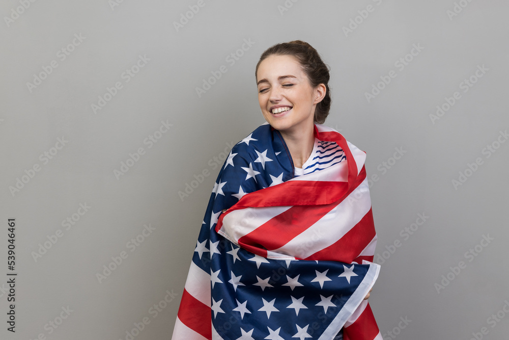 Portrait of delighted optimistic woman in striped T-shirt standing wrapped in USA flag, expressing happiness, posing with closed eyes and toothy smile. Indoor studio shot isolated on gray background. Portrait of delighted optimistic woman in striped T-shirt standing wrapped in USA flag, expressing happiness, posing with closed eyes and toothy smile. Indoor studio shot isolated on gray background.