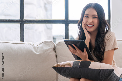 Canvas Print Freelance asian woman surfing browser with tablet sitting with pillow on sofa at living room