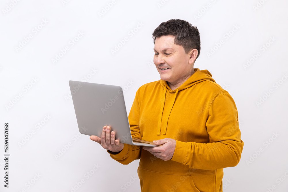 Attractive middle aged man working on laptop typing email or chatting in social network, happy face expression, wearing urban style hoodie. Indoor studio shot isolated on white background. Attractive middle aged man working on laptop typing email or chatting in social network, happy face expression, wearing urban style hoodie. Indoor studio shot isolated on white background.